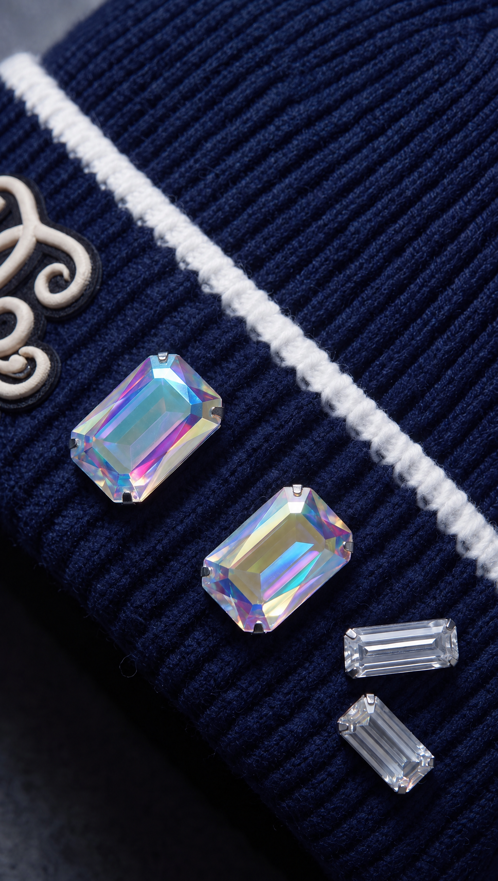 Gros plan du revers du bonnet The King Crown bleu marine : monogramme Dellerium en cordon ivoire, cristaux taille émeraude à reflets aurore boréale et ligne contrastée